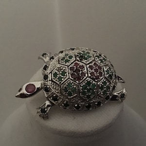 May include: A silver turtle brooch with a detailed design. The turtle's shell is decorated with green, red, and white gemstones.