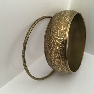 May include: Two gold-toned metal bangles. One bangle has a simple, textured design. The other bangle has an intricate floral design.