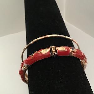 May include: Two gold bangles, one with red enamel and floral designs, the other with a simple gold finish.