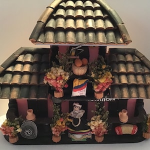 May include: A two-story wooden model of a Colombian cafe with a green tiled roof, a pink facade, and a sign that reads "Cafe de Colombia". The model is decorated with dried flowers, a small doll in traditional Colombian dress, and a small accordion.