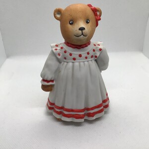 May include: A porcelain figurine of a brown teddy bear wearing a white dress with red polka dots and red trim. The bear has a red bow on its head.