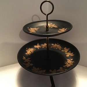 May include: Two-tiered serving stand with black ceramic plates and gold floral designs. The stand has a metal base and a gold handle.
