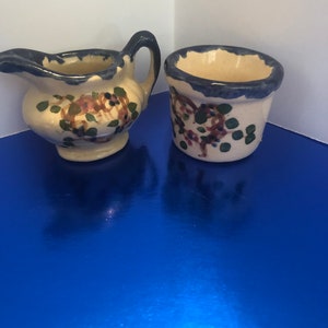 May include: A set of two small, hand-painted ceramic pieces. The creamer is white with blue trim and a floral design. The small cup is also white with blue trim and a floral design.