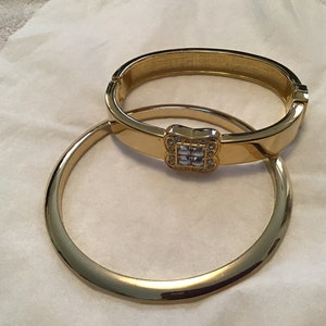 May include: Two gold bangle bracelets, one with a square-shaped clear crystal embellishment.