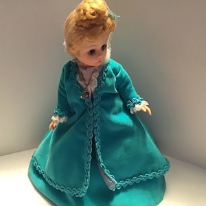 May include: A vintage porcelain doll with blonde hair wearing a teal blue dress with white lace trim and a matching jacket. The doll has a necklace with a pendant.