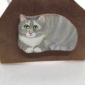 May include: A gray and white cat with green eyes is painted on a brown paper background. The cat is curled up in a sleeping position.