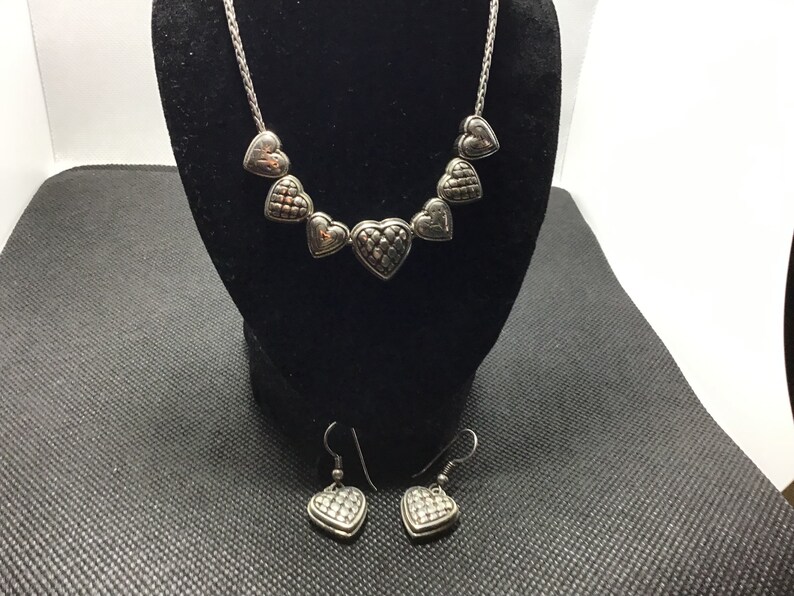 Puede incluir: Un collar y pendientes de plata con una cadena y colgantes en forma de coraz&oacute;n. Los corazones tienen un dise&ntilde;o texturizado y acolchado.