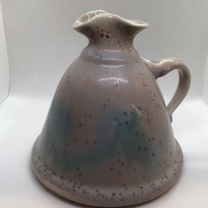 May include: A ceramic pitcher with a speckled glaze in shades of purple, blue, and green. The pitcher has a wide, rounded base and a flared spout.