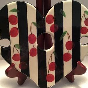 May include: A heart-shaped ceramic wine bottle holder with a black and white striped design and red cherries painted on it. The holder has a cut-out circle in the center for the bottle neck.
