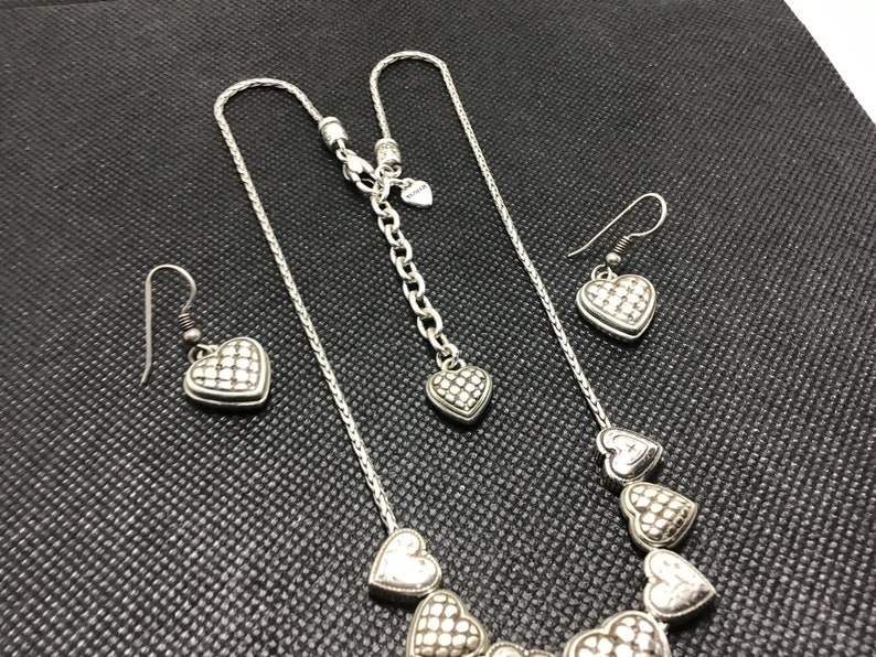 Puede incluir: Un collar de plata con un colgante de coraz&oacute;n y pendientes a juego. El collar tiene una cadena con m&uacute;ltiples colgantes de coraz&oacute;n. Los pendientes son simples y tienen un solo colgante de coraz&oacute;n.