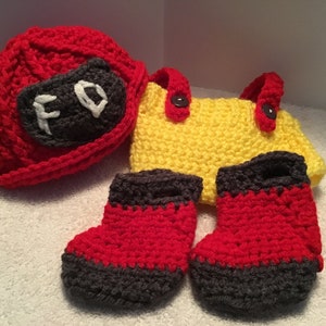 May include: A handmade crochet baby set in red, yellow, and gray. The set includes a firefighter hat with "FD" on it, a yellow bag with red handles, and a pair of red and gray booties. The items are made of soft yarn.