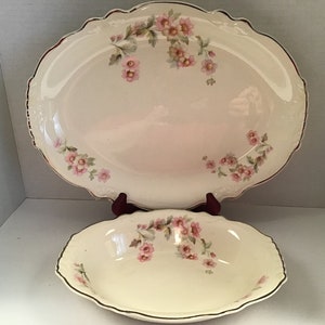 May include: A set of two vintage oval-shaped ceramic serving dishes with a floral design. The larger dish is a platter with a scalloped edge and the smaller dish is a bowl. Both dishes feature a white background with pink flowers and green leaves.