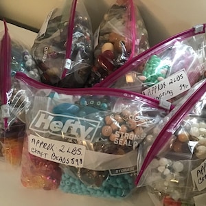 May include: Five clear plastic bags filled with various colorful beads. The bags are labeled with handwritten notes indicating the type and weight of the beads. The bags are labeled "APPROX 2 LBS. CRAFT BEADS $98", "APPROX 2 LBS. CRAFTING 196", "APPROX 2 LBS. CRAFTING 196", and "APPROX 2 LBS. CRAFTING 196".