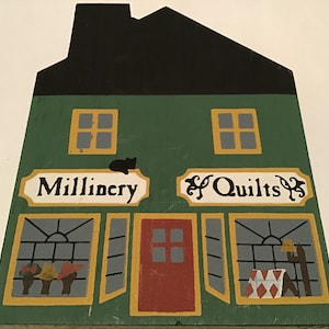 May include: A green wooden building with a black roof. The building has two windows and two doors. The sign above the left door says "Millinery" and the sign above the right door says "& Quilts".