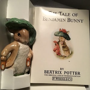 May include: A porcelain figurine of Peter Rabbit wearing a green hat and holding a carrot. The figurine is next to a book titled "The Tale of Benjamin Bunny" by Beatrix Potter.