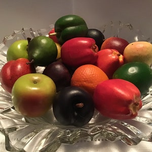 May include: A clear, decorative glass bowl filled with a variety of colorful artificial fruits. The fruits include green apples, red plums, oranges, and peaches. The bowl has a starburst design.
