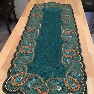 May include: Teal beaded table runner with a gold and teal paisley design. The runner has a scalloped edge.