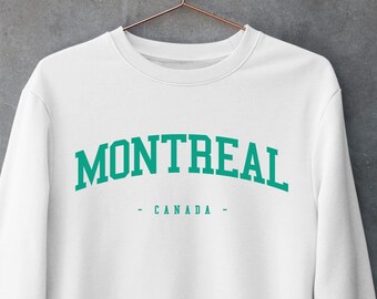 May include: A white crewneck sweatshirt featuring the word MONTREAL in a large, arched, teal-colored serif font, with the word CANADA printed in a smaller, straight teal font below it, all displayed on a garment hanging from a copper hanger.
