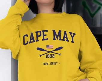 May include: A mustard yellow crewneck sweatshirt featuring the dark blue text CAPE MAY, a small American flag, crossed oars, the year 1692, and the text NEW JERSEY. The sweatshirt has long sleeves with ribbed cuffs and a ribbed hem.