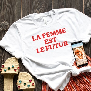 May include: White t-shirt with the text "LA FEMME EST LE FUTUR" in red lettering. The shirt is paired with orange and white striped pants, a pair of brown sandals with colorful jewels, and a clear pair of glasses. A cell phone with a picture of a puppy is on the shirt.