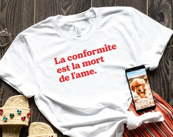 May include: White t-shirt with the red text "La conformité est la mort de l'âme." A smartphone displays a golden retriever puppy image. The shirt is paired with orange striped shorts and embellished sandals on a dark wood surface.