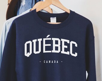 Puede incluir: Sudadera azul marino con la palabra "QUÉBEC" en letras blancas, con "CANADA" debajo. La sudadera está colgada en una percha de madera. Un sombrero marrón y una camisa vaquera son visibles en el fondo.