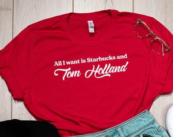 May include: Red t-shirt with white text that reads "All I want is Starbucks and Tom Holland". The shirt is laid out on a wooden surface with a black baseball cap, blue denim shorts, a bracelet, and white sneakers.