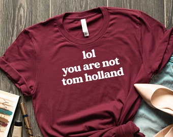 May include: A maroon t-shirt with white text that reads "lol you are not tom holland". The shirt is laid out on a wooden surface with a pair of jeans, a pair of high heels, and makeup.