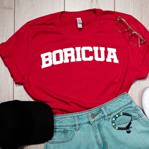 May include: Red t-shirt with white text that says "BORICUA" on it. The shirt is laid out on a wooden surface with a black baseball cap, blue denim shorts, white trainers, and clear glasses.