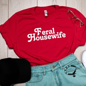 May include: Red t-shirt with white text that says "Feral Housewife". The shirt is laid out on a wooden surface with a black baseball cap, blue denim shorts, a bracelet, and white sneakers.