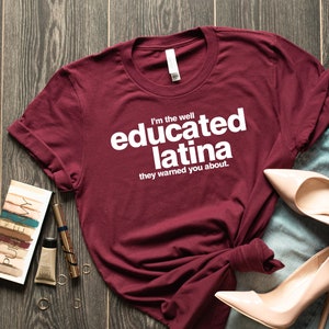 Può includere: Una maglietta bordeaux con la scritta "I'm the well educated latina they warned you about." è abbinata a jeans chiari, tacchi nude e trucco su una superficie di legno. Gli accessori includono una collana e anelli.