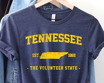 Puede incluir: Una camiseta azul marino con un gráfico amarillo del estado de Tennessee, el texto "Tennessee", "Est. 1868", y "The Volunteer State".