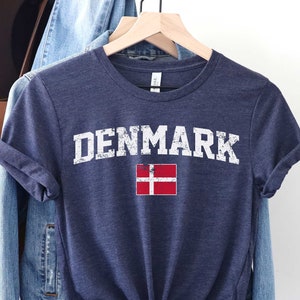 K&ouml;nnte beinhalten: Ein marineblaues T-Shirt mit dem Wort "DENMARK" in wei&szlig;er, abgenutzter Schrift und einem Grafikdruck der d&auml;nischen Flagge in Rot und Wei&szlig;.