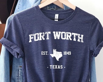May include: A navy blue t-shirt with the text "FORT WORTH" in white, along with "EST. 1849" and a Texas outline. The shirt is knotted at the waist and hangs with a denim jacket and a brown hat.