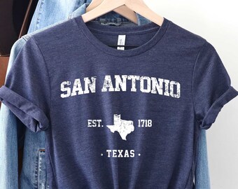 May include: A navy blue t-shirt with a white distressed graphic that says "SAN ANTONIO EST. 1718 . TEXAS."