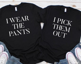 Könnte beinhalten: Zwei schwarze T-Shirts mit weißem Text. Ein Shirt sagt "Ich trage die Hosen" und das andere sagt "Ich wähle sie aus".