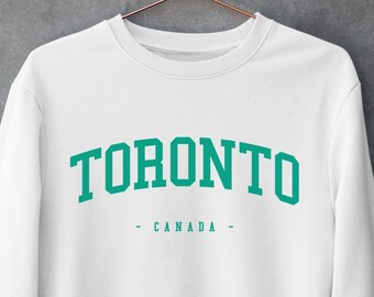 Puede incluir: Una sudadera blanca de cuello redondo que presenta la palabra TORONTO en una fuente verde grande y arqueada de estilo universitario, con la palabra CANADA centrada debajo en una fuente verde más pequeña, flanqueada por dos guiones verdes.