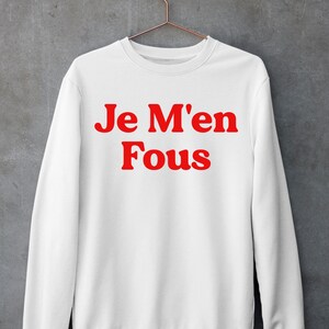 Puede incluir: Sudadera blanca con texto rojo que dice "Je M'en Fous".