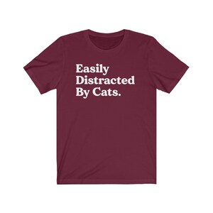 Może przedstawiać: Bordowy T-shirt z kr&oacute;tkim rękawem z białym napisem "Easily Distracted By Cats."
