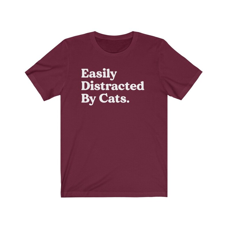 Może przedstawiać: Bordowy T-shirt z kr&oacute;tkim rękawem z białym napisem "Easily Distracted By Cats."