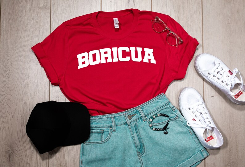 May include: Red t-shirt with white text that says "BORICUA" on it. The shirt is laid out on a wooden surface with a black baseball cap, blue denim shorts, white trainers, and clear glasses.