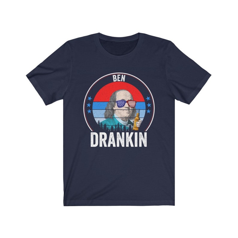 Puede incluir: Una camiseta azul marino con un gr&aacute;fico de estilo vintage de Benjamin Franklin con gafas de sol y sosteniendo una cerveza. El gr&aacute;fico est&aacute; rodeado de un c&iacute;rculo rojo, blanco y azul con estrellas y el texto "BEN DRANKIN".