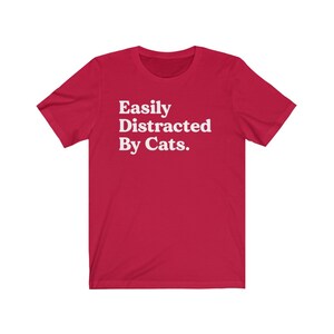 Może przedstawiać: Czerwona koszulka z kr&oacute;tkim rękawem z białym napisem "Easily Distracted By Cats."
