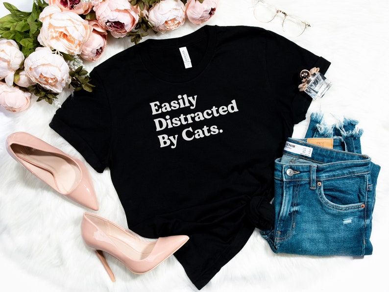 Może przedstawiać: Czarny T-shirt z białym napisem "Easily Distracted By Cats."