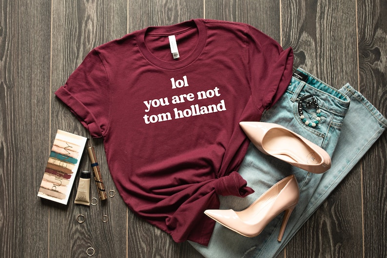 May include: A maroon t-shirt with white text that reads "lol you are not tom holland". The shirt is laid out on a wooden surface with a pair of jeans, a pair of high heels, and makeup.