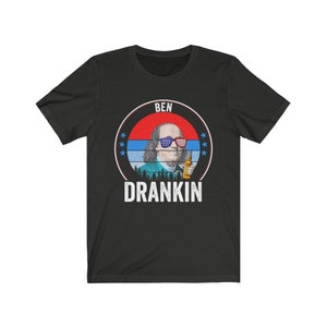 Puede incluir: Camiseta negra con un gr&aacute;fico de Benjamin Franklin con gafas de sol rojo, blanco y azul y sosteniendo una cerveza. El gr&aacute;fico est&aacute; rodeado de un c&iacute;rculo rojo, blanco y azul con estrellas y el texto "BEN DRANKIN".