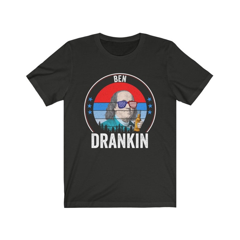 Puede incluir: Camiseta negra con un gr&aacute;fico de Benjamin Franklin con gafas de sol rojo, blanco y azul y sosteniendo una cerveza. El gr&aacute;fico est&aacute; rodeado de un c&iacute;rculo rojo, blanco y azul con estrellas y el texto "BEN DRANKIN".