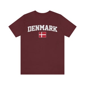 K&ouml;nnte beinhalten: Ein maroonfarbenes T-Shirt mit einem wei&szlig;en, abgenutzten Aufdruck des Wortes "DENMARK" und der d&auml;nischen Flagge.