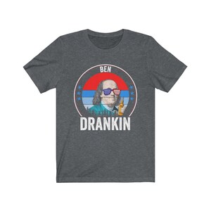 Puede incluir: Una camiseta gris oscuro jaspeada con un gr&aacute;fico de estilo vintage de Benjamin Franklin con gafas de sol y sosteniendo una cerveza. El gr&aacute;fico est&aacute; rodeado de estrellas y el texto "BEN DRANKIN".