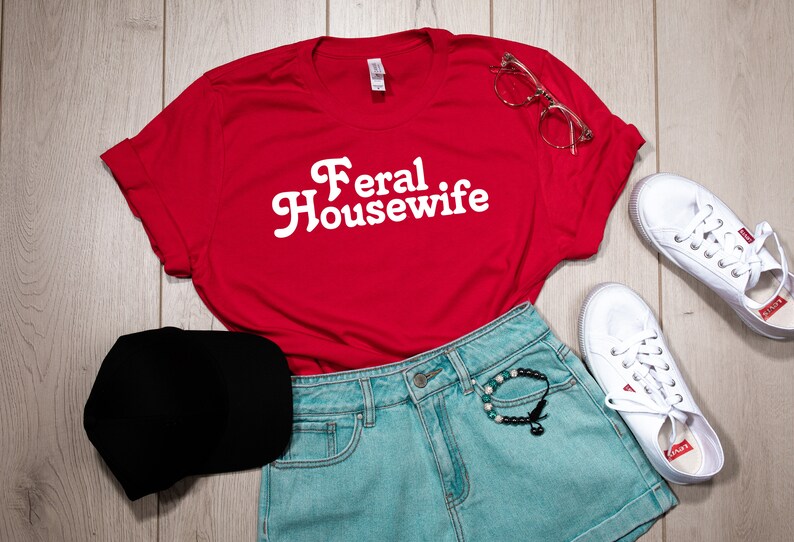 May include: Red t-shirt with white text that says "Feral Housewife". The shirt is laid out on a wooden surface with a black baseball cap, blue denim shorts, a bracelet, and white sneakers.